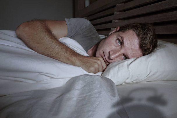 restless worried young attractive man awake at night lying on bed sleepless having eyes opened depressed suffering insomnia sleeping disorder with classic alarm clock shadow in problem concept - Foto, afbeelding