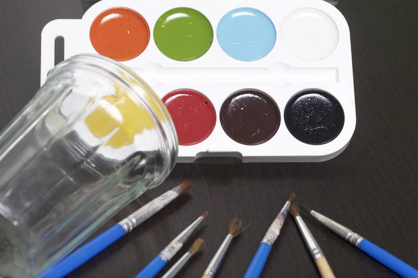 Watercolors and brushes on a dark surface. Nearby is a glass for water. Subjects for creativity. - Photo, image