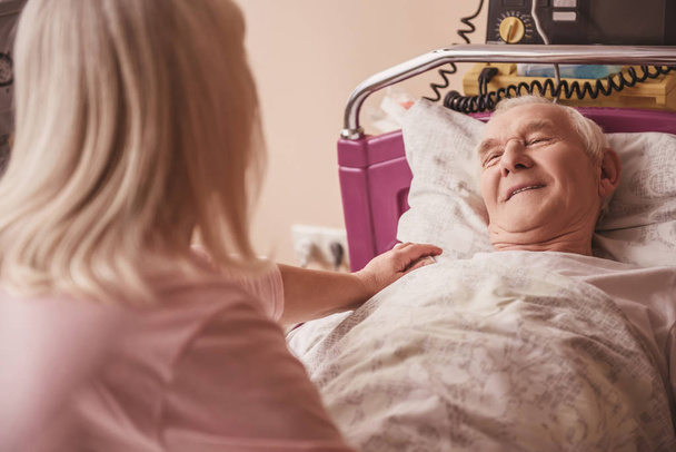 Cheerful old man is looking at his wife and smiling while lying in bed in the hospital ward - Foto, afbeelding