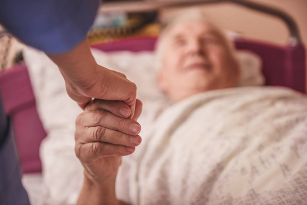 geschnittenes Bild eines alten Mannes, der die Krankenschwester ansieht, ihre Hand hält und lächelt, während er im Krankenhausbett liegt - Foto, Bild