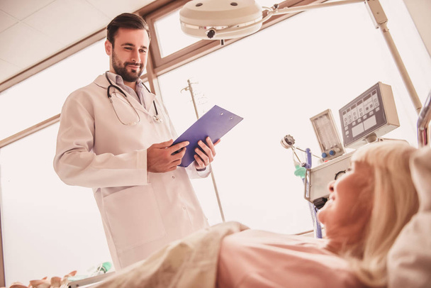 Przystojny lekarz ogląda jego żona, leżąc w łóżku w oddziale szpitala i uśmiechając się - Zdjęcie, obraz