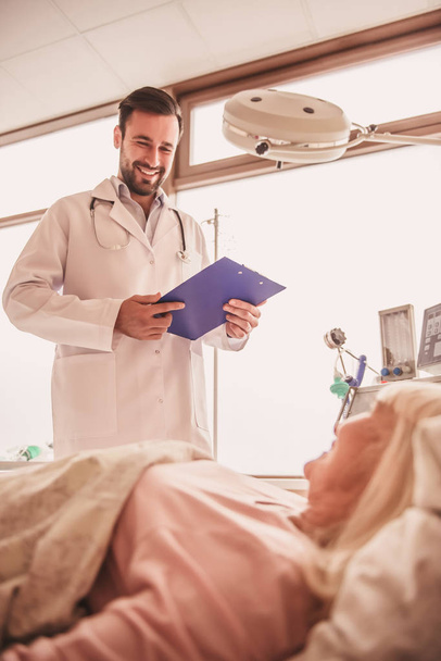 Médico bonito está olhando para sua esposa deitada na cama na enfermaria do hospital e sorrindo
 - Foto, Imagem