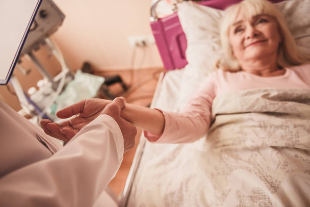 病棟でベッドに横たわっている間陽気な老婦人は笑っている、医師は彼女の脈拍数を取っています。 - 写真・画像