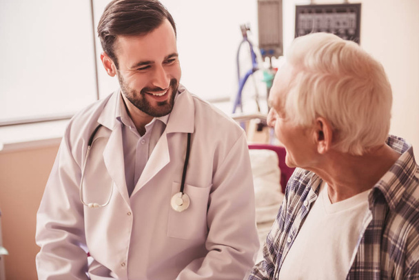 Guapo médico está hablando con un hombre viejo y sonriendo mientras está sentado en la sala de hospital
 - Foto, Imagen