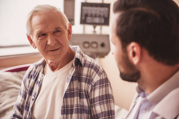 Handsome old man is talking with a doctor while sitting in hospital ward - Foto, imagen
