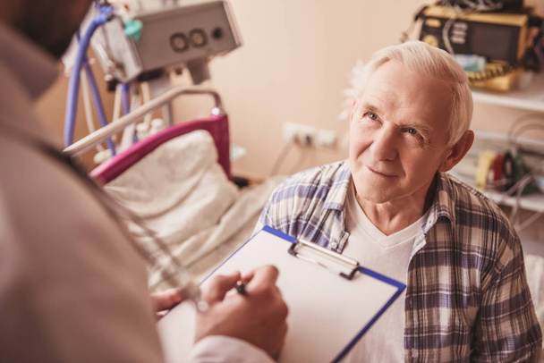 Hombre viejo guapo está hablando con un médico mientras está sentado en la sala de hospital
 - Foto, Imagen