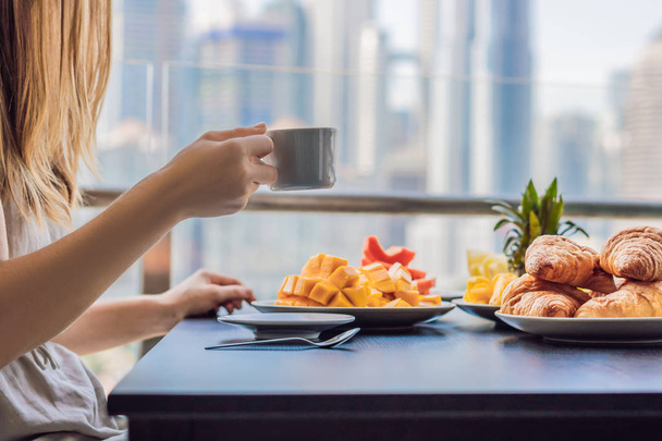Young woman having breakfast on balcony. Breakfast table with coffee. fruits and bread croissant on balcony against backdrop of big city - Фото, зображення
