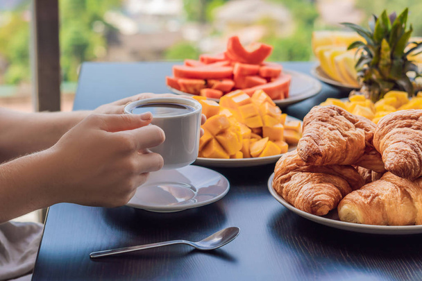 Young woman having breakfast on balcony. Breakfast table with coffee. fruits and bread croissant on balcony against backdrop of big city - Foto, Imagen
