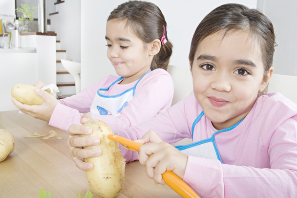 Две одинаковые сестры-близнецы чистят картошку на домашнем кухонном столе
. - Фото, изображение