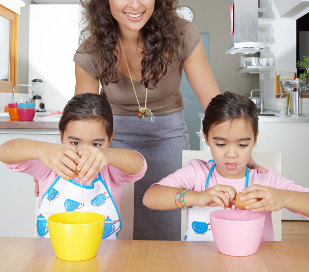 若い双子姉妹の母それらの横にある自宅の台所で笑みを浮かべて立って中ボウルに卵を開く. - 写真・画像