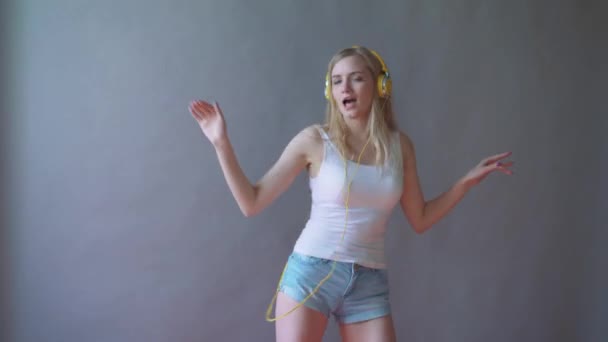 Young beautiful woman on a gray background, listening to music in headphones on the phone, dancing - Footage, Video