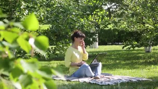 Girl freelancer works in the open air.The brunette sits on the grass in the park and talks on the phone. On her lap she has a computer. - Кадры, видео