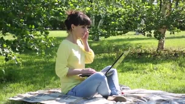 Girl freelancer works in the open air.The brunette sits on the grass in the park and talks on the phone. On her lap she has a computer. - Filmagem, Vídeo