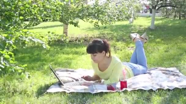 Dívka na volné noze pracuje pod širým nebem. Ta bruneta sedí na trávě v parku a pracuje s přenosným počítačem. Tiskne na klávesnici zprávu na sociální síti na internetu. - Záběry, video