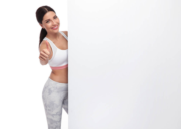 Γυμναστήριο ελκυστικό κορίτσι κάνει εντάξει σημάδι κρατώντας πινακίδα. Στέκεται σε λευκό φόντο, χαμογελώντας, ενώ φοράει ένα ζευγάρι άσπρα κολάν και ένα άσπρο πάνω - Φωτογραφία, εικόνα