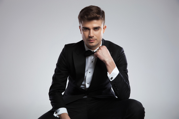 portrait of seated young businessman in tuxedo fixing his black bowtie on light grey background - Photo, Image