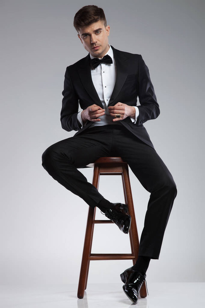 verführerischer Geschäftsmann, der auf einem Holzstuhl sitzt und seine schwarze Anzugjacke auf hellgrauem Hintergrund zuknöpft - Foto, Bild
