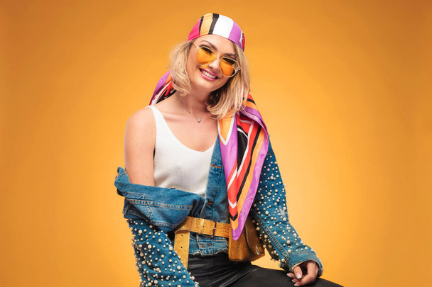 portrait of smiling blonde woman wearing a colorful rock outfit sitting on yellow background - Photo, Image