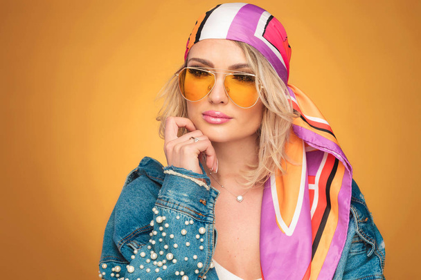 portrait of pensive rock woman wearing a colored headscarf and yellow sunglasses looking to side on orange background - Photo, Image