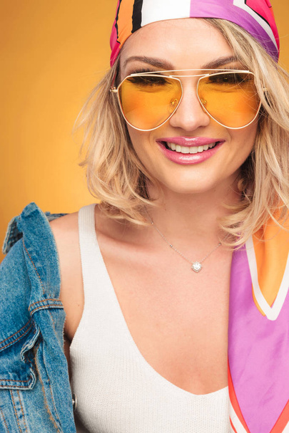 крупним планом блондинка з кольоровим головним убором і жовтими сонцезахисними окулярами, посміхаючись на жовтому фоні
 - Фото, зображення