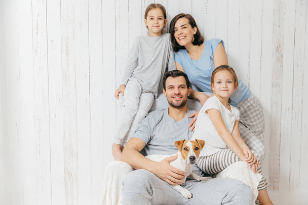 Horizontal shot of friendly family pose together against white background: two little sisters, father, mother and their pet. Happy parents and their female children. Family of four. Parenthood - Photo, Image