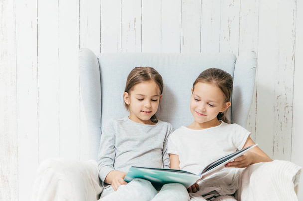 Two little adorable sisters sit in sofa, read interesting book, sit in comfortable sofa, enjoy togetherness. Best friends female children hold book, view pictures and read hearders, have spare time - Fotó, kép