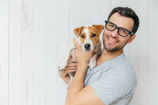 Horizontal portrait of handsome cheerful man, wears eyeglasses, holds jack russell terrirer, has glad expression, poses against white wooden wall with blank copy space. Animals and friendship - Photo, Image
