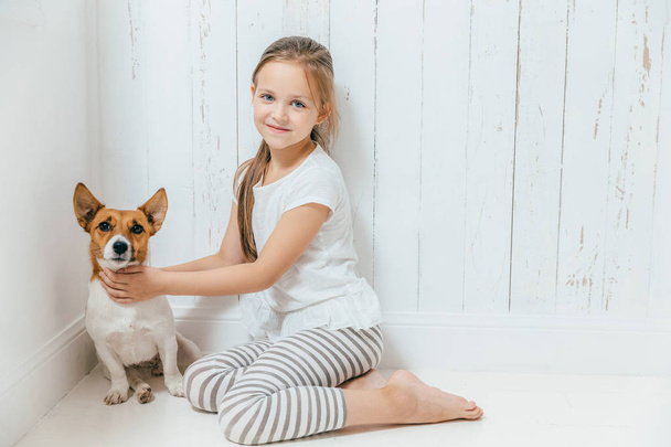Υπέροχο μικρό θηλυκό παιδί παίζει με το σκύλο της στο λευκό δωμάτιο, κάθονται στο πάτωμα, έχουν καλή σχέση, αγκαλιές αγαπημένο κατοικίδιο. Μικρή μαθήτρια αρέσει ζώα, περνά το ελεύθερο χρόνο σας στο σπίτι. Παιδική ηλικία - Φωτογραφία, εικόνα