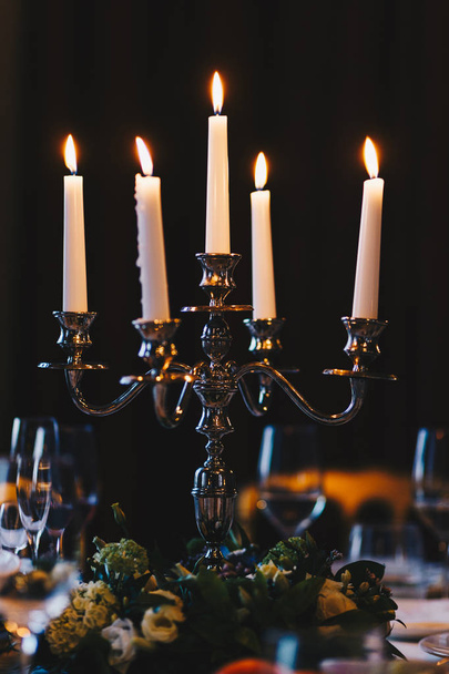 Colpo verticale di bellissimo candeliere con cinque candele bianche accese sullo sfondo scuro o nella stanza. Candele antiche su tavola decorata
 - Foto, immagini