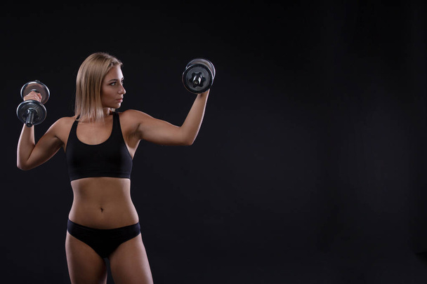 Brutal athletic woman pumping up muscles with dumbbells on black background. Copy space - Photo, image