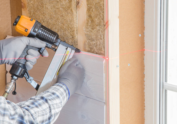 Работник выполняет отделочные работы стен с белой деревянной доской, используя уровень лазерной линии. Строительство теплоизоляционного экологически-деревянного каркасного дома из древесного волокна
 - Фото, изображение
