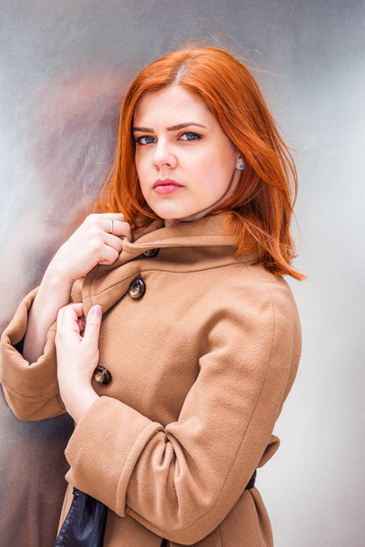 Portrait of Beautiful Young American Woman in New York, wearing long brown woolen overcoat, hands touching coat in front, standing by silver wall in winter day, looking at yo - Fotografie, Obrázek