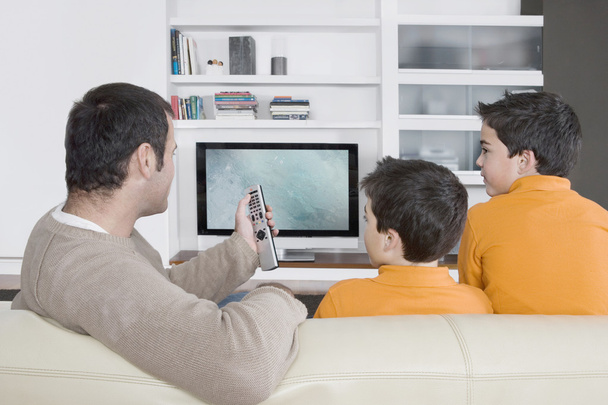 Отец с братьями-близнецами смотрит телевизор дома, используя пульт дистанционного управления
. - Фото, изображение