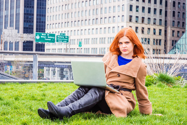 junge amerikanische Geschäftsfrau auf Reisen, arbeitet in New York, trägt einen langen braunen Wollmantel, lange schwarze Lederstiefel, sitzt auf dem grünen Rasen im Geschäftsviertel, arbeitet am Laptop, denkt nach - Foto, Bild
