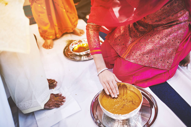 The bride standing on the white fabric and the women prepraing the turmeric (haldi) oil mixed with milk to pasting on the bride's feet and body, the traditional rituals of indian marriage ceremony - 写真・画像