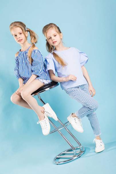 Mädchen-Zwillinge in hellblauer Kleidung posieren in der Nähe eines Barhockers auf blauem Hintergrund. - Foto, Bild