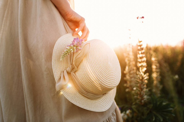 Woman holding straw hat with a ribbon on flower field background. Warm sunset light beams - Photo, Image