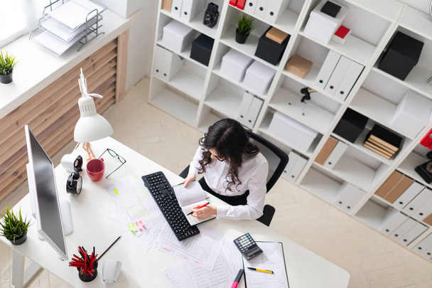 Ein junges Mädchen sitzt im Büro am Computertisch, hält einen Stift in der Hand und blickt auf den Notizblock. - Foto, Bild