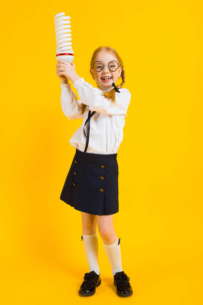 Meisje met rode pigtails op een gele achtergrond. Een charmante meisje in ronde transparante glazen houdt een gloeilamp in haar handen. - Foto, afbeelding