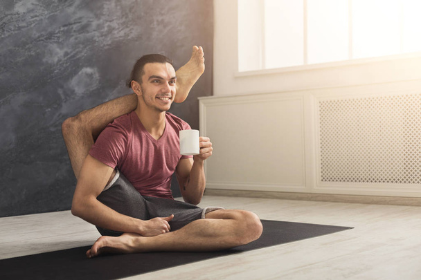 Young sporty man in flexible yoga pose drinking tea, sitting on mat at gym and practicing twisting exercise, copy space - Photo, Image