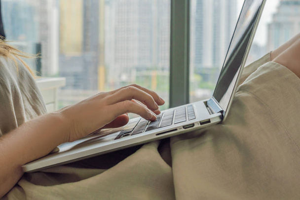 Young woman is working on laptop on balcony overlooking skyscrapers. Freelancer, remote work, work from home. - Foto, imagen