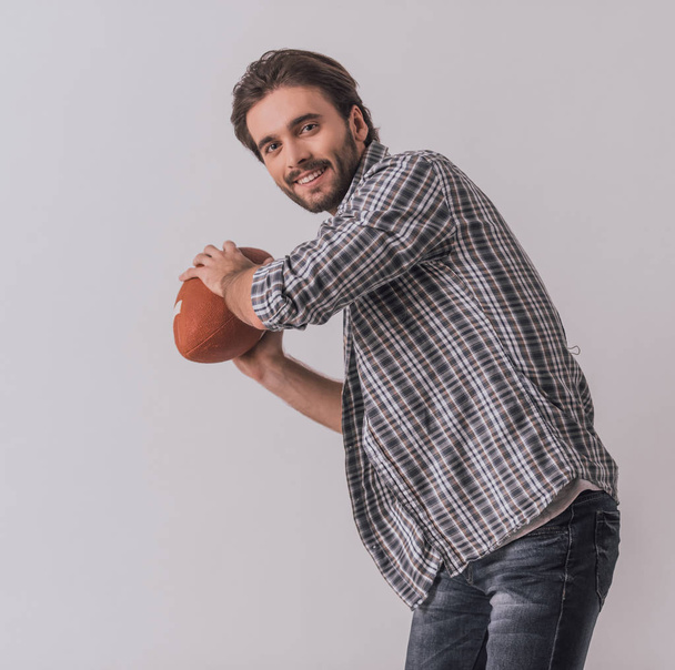 Handsome bearded man in casual clothes is throwing a football ball, looking at camera and smiling, on light background - Фото, зображення