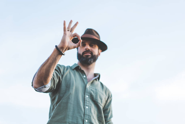 Handsome happy bearded hipster looking at camera through his fingers in ok gesture. People Watching concept. - Foto, Bild