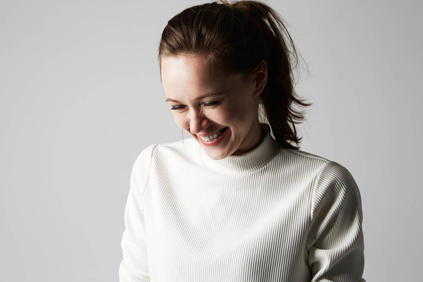 Fashion photo of a beautiful smiling brunette woman in a white roll-neck sweater posing over gray empty background. Fashionable photo shot. - 写真・画像