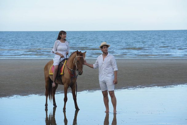 Romantic Couple With horse riding At the beach in the afternoon On a sweet holiday.Young couple Exercise and jump at the beach in the early morning sunrise. With beautiful yellow sky - Photo, Image