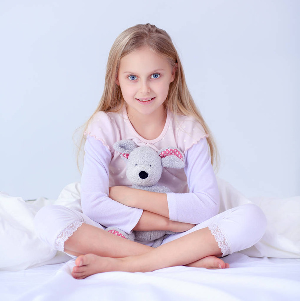 Little girl in the bedroom is sitting on the bed. Little girl is wearing a pajamas and sitting in bed - Foto, imagen