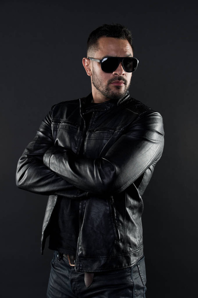 Confident man stand with arms folded. Bearded man in trendy sunglasses. Fashion model in leather jacket and jeans. Fashion and style. Confidence with sexuality and charisma - Φωτογραφία, εικόνα