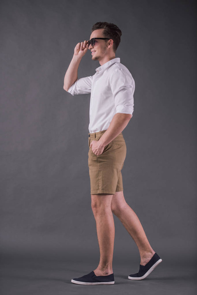 Full-length, back view image of handsome young man in shirt, shorts and sun glasses smiling while walking on gray background - Fotografie, Obrázek