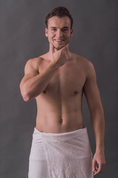 Handsome young man wrapped in towel is touching his shaved chin, looking at camera and smiling, on gray background - Fotó, kép