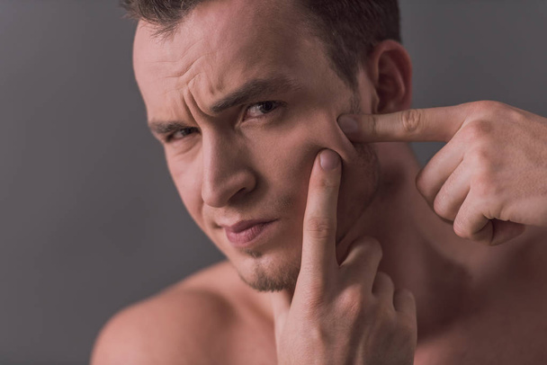Handsome young man with bare torso is squeezing pimples, on gray background - Photo, image